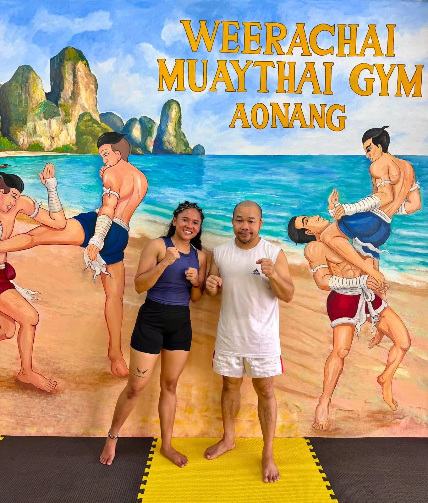 Two people posing in front of the gym mural