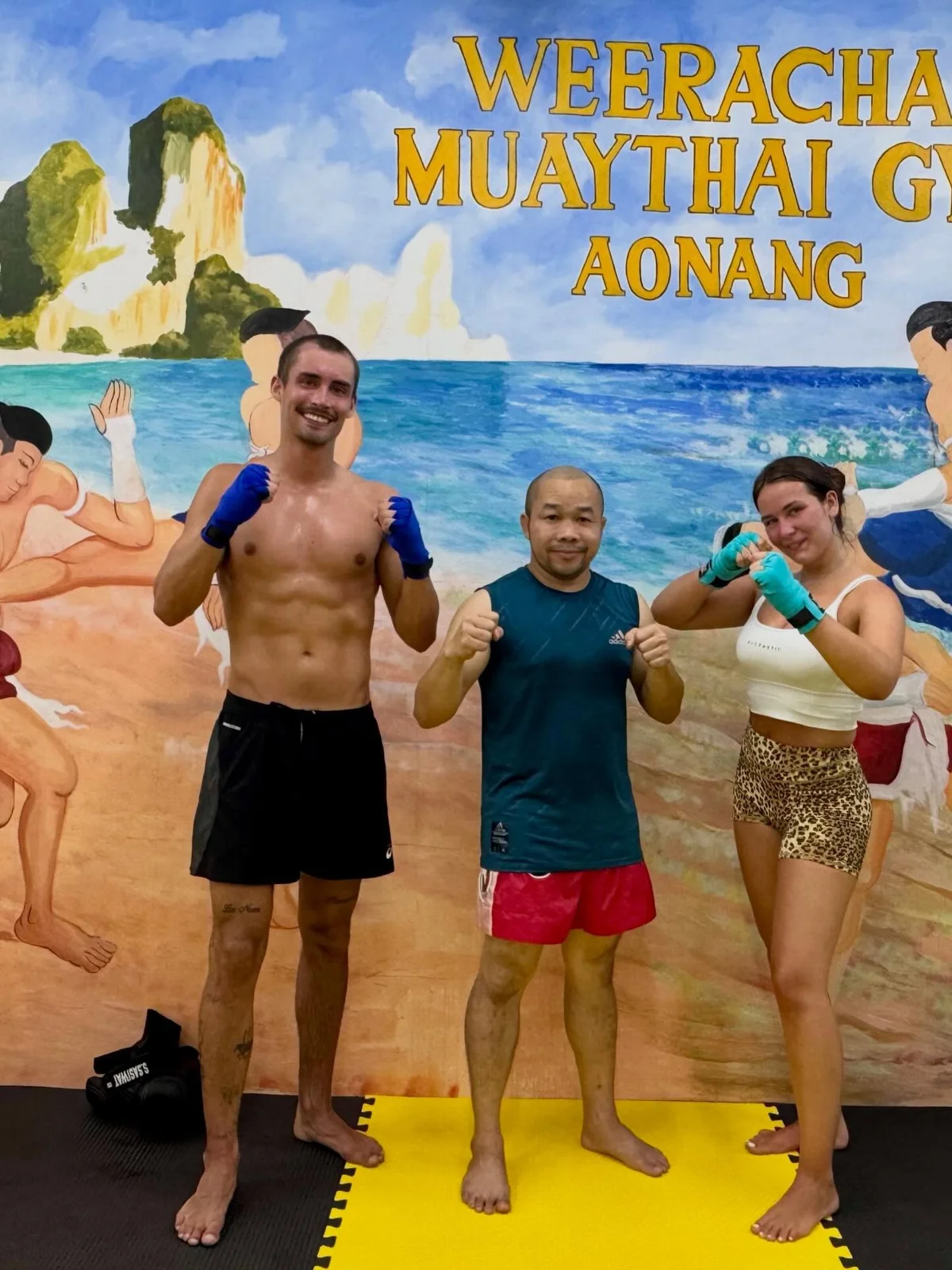 Three people posing with boxing gloves in front of the Weerachai Muaythai Gym mural